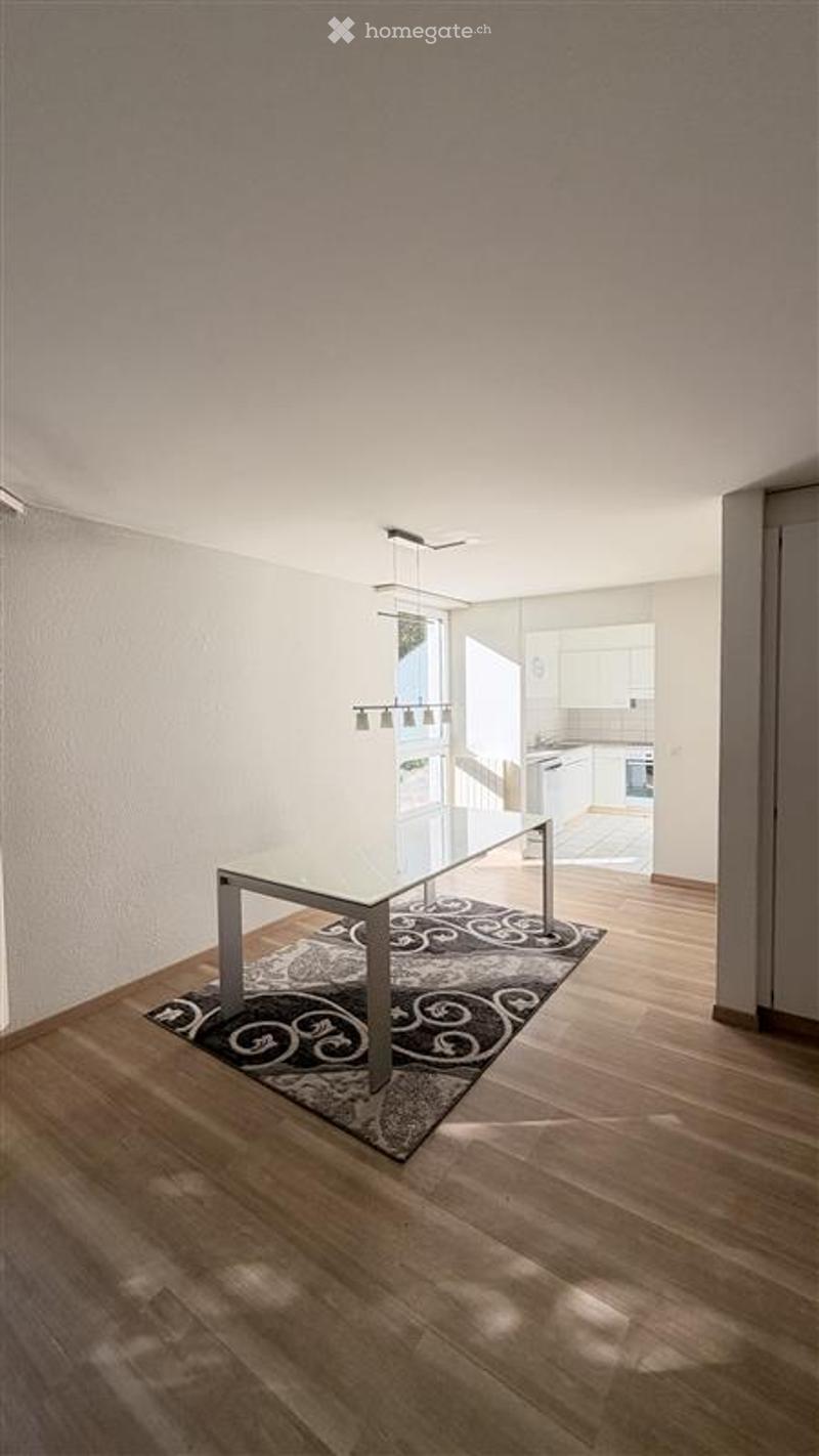Bright, minimalist dining area with a sleek glass dining table, a patterned rug, and modern pendant lighting. The room features light wooden flooring and white walls, with an open view to a contemporary kitchen in the background.