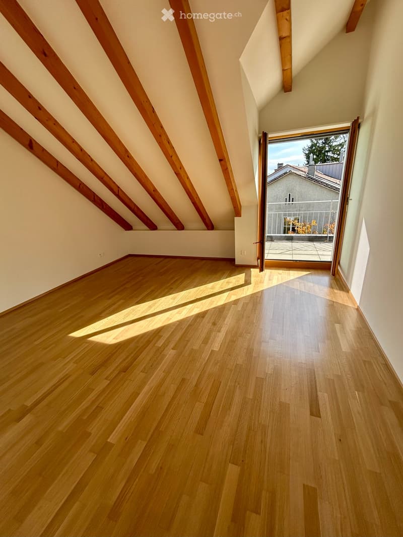 Empty attic space with wooden floors, exposed wooden beams, and a large window opening to a balcony area.
