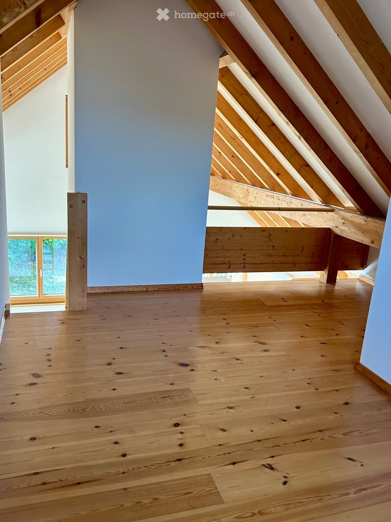 An empty attic space with wooden flooring, exposed wooden beams, and a slanted ceiling. The walls are painted in light blue and white, with a window on one side allowing natural light to enter.