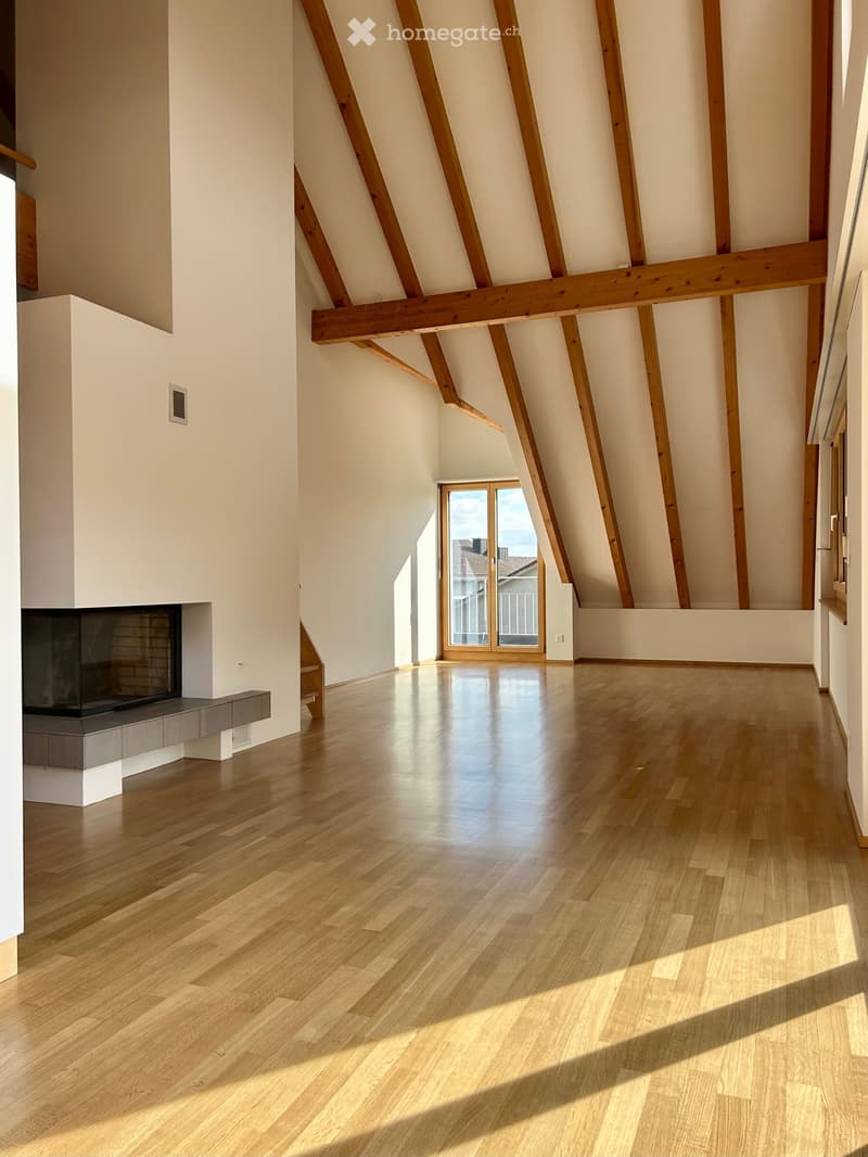 Bright, open attic space with wooden beams, polished hardwood floors, and a modern fireplace on the left side. Large windows and a glass door lead to a balcony.