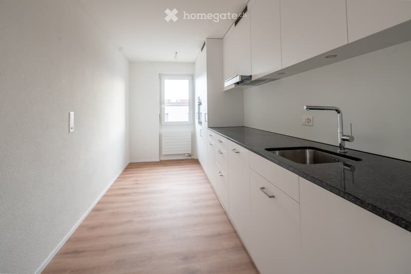 Modern kitchen with white cabinets, dark countertop, and stainless steel sink and faucet. Wooden flooring and a window at the end.
