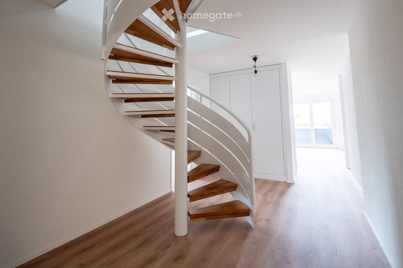 Spiral staircase with wooden steps and white railing, leading to a bright hallway with light wooden flooring and white walls.