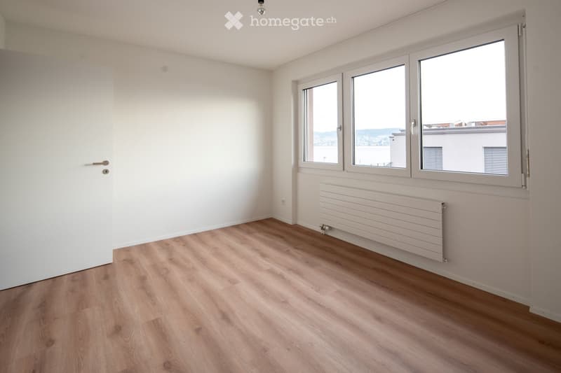 Empty room with light wooden flooring, white walls, large windows with a view, and a radiator under the windows.