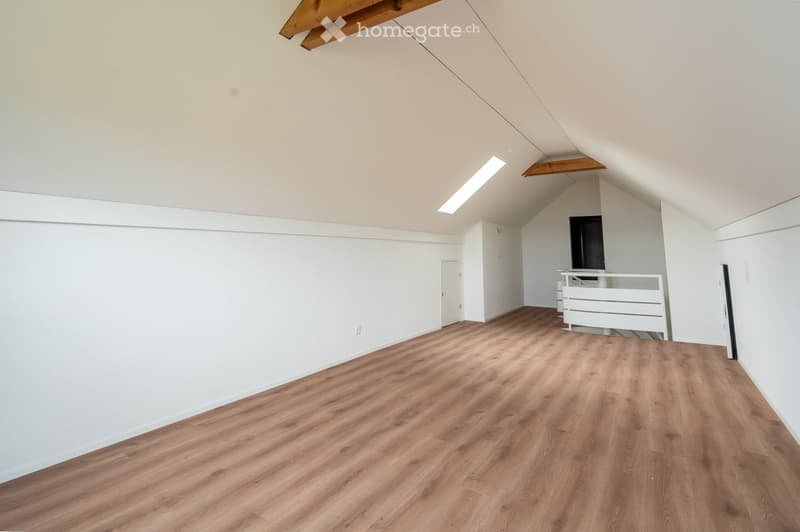 Empty attic room with wooden flooring, white walls, and exposed wooden beams on the ceiling. Features a small white railing and a closed door leading to another area.