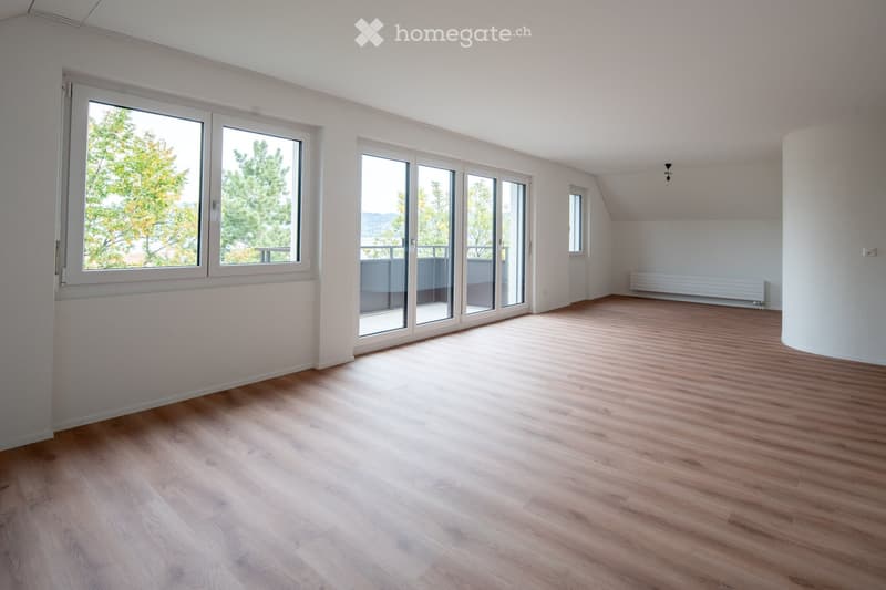 Empty room with wooden flooring, large windows, and sliding glass doors leading to a balcony. The walls are white, and there is a radiator on one side.