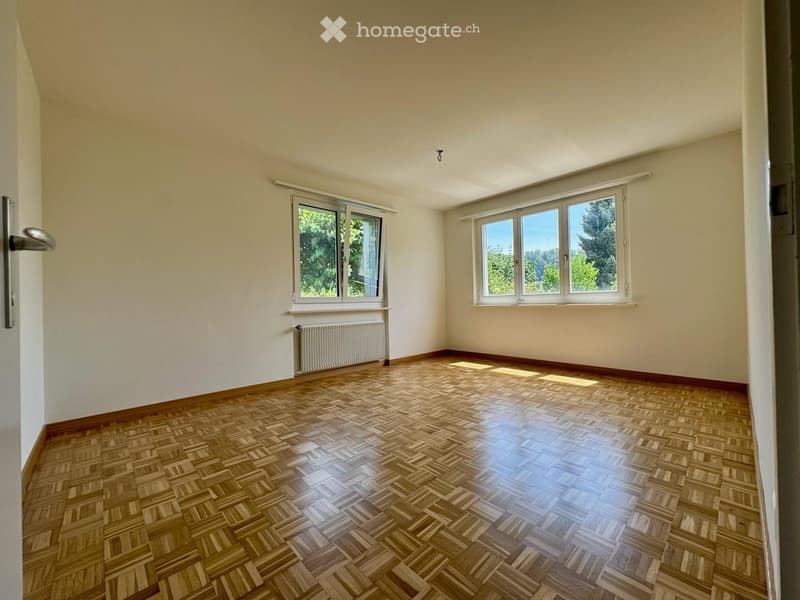 Empty room with wooden herringbone flooring, white walls, large windows, and a radiator. No furniture or equipment present.