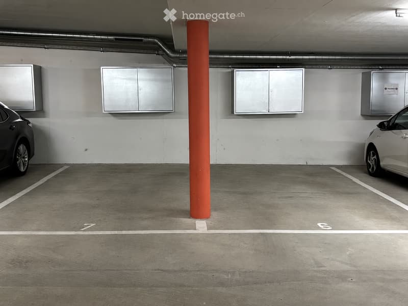 Underground parking area with concrete floor, white walls, and two parked cars. Features a red support pillar and several metal cabinets mounted on the wall.