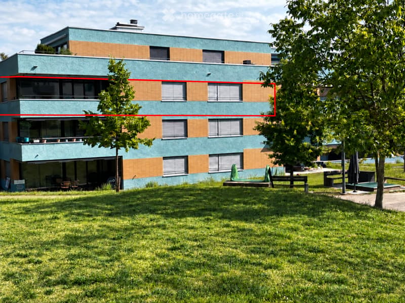 Modern two-story building with light blue facade and brown accents, featuring large windows and balconies, surrounded by green lawn and trees.