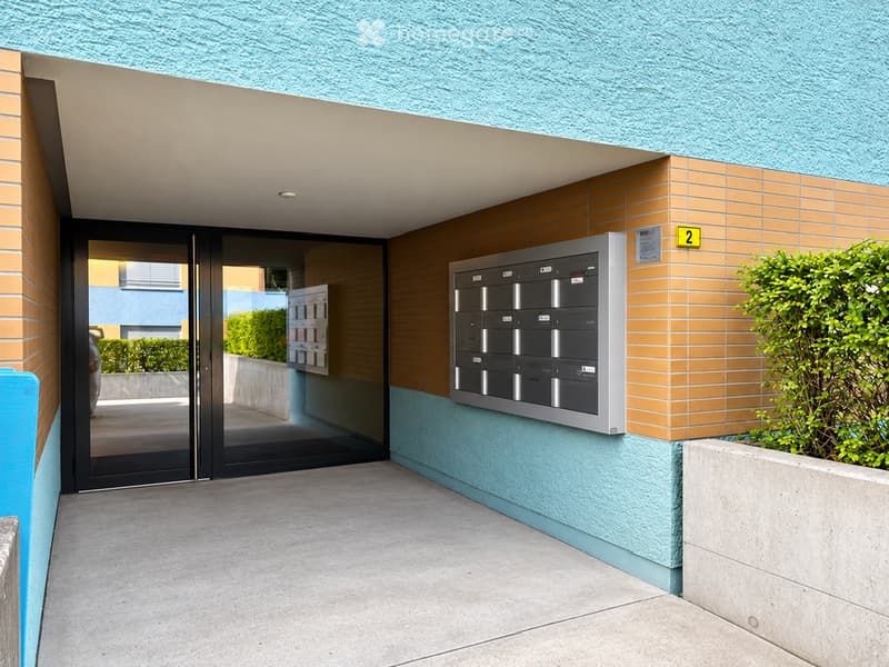 Modern building entrance with mailboxes, glass doors, and a small garden area.