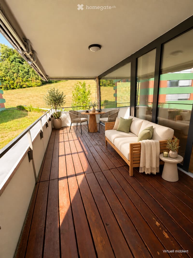 A modern wooden balcony with a white sofa, small round table, chairs, potted plants, and candles, overlooking a green field.