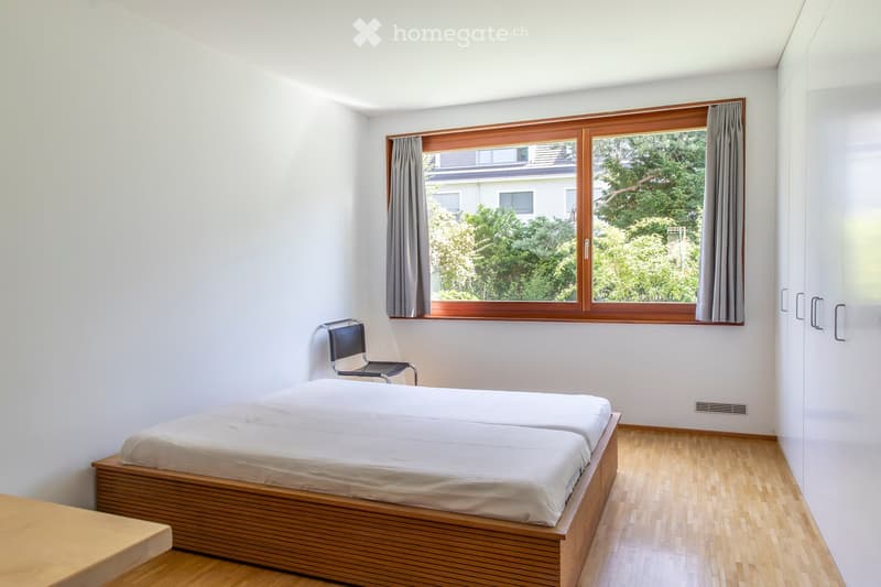 Minimalist bedroom with a single bed, a chair, and large windows with wooden frames and grey curtains. The room features light-colored walls and a wooden floor.