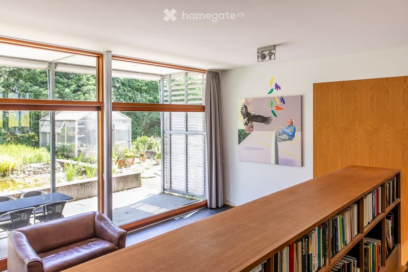 Bright living room with large windows overlooking a garden, featuring a leather sofa, wooden bookshelf, dining table, and chairs.