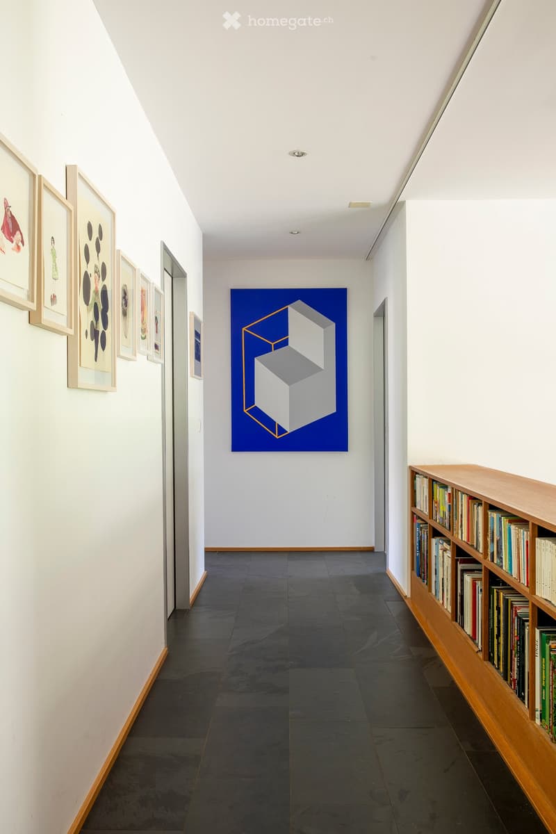 Modern hallway with white walls, dark tiled floor, wooden shelving unit filled with books, and framed artwork on the walls.
