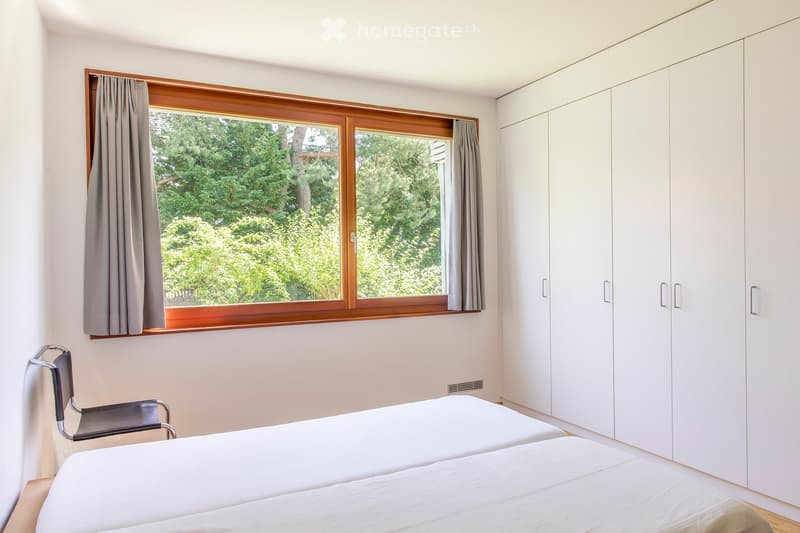 Bright bedroom with two single beds, a chair, and a large built-in wardrobe. Large windows with wooden frames and grey curtains offer a view of greenery outside.