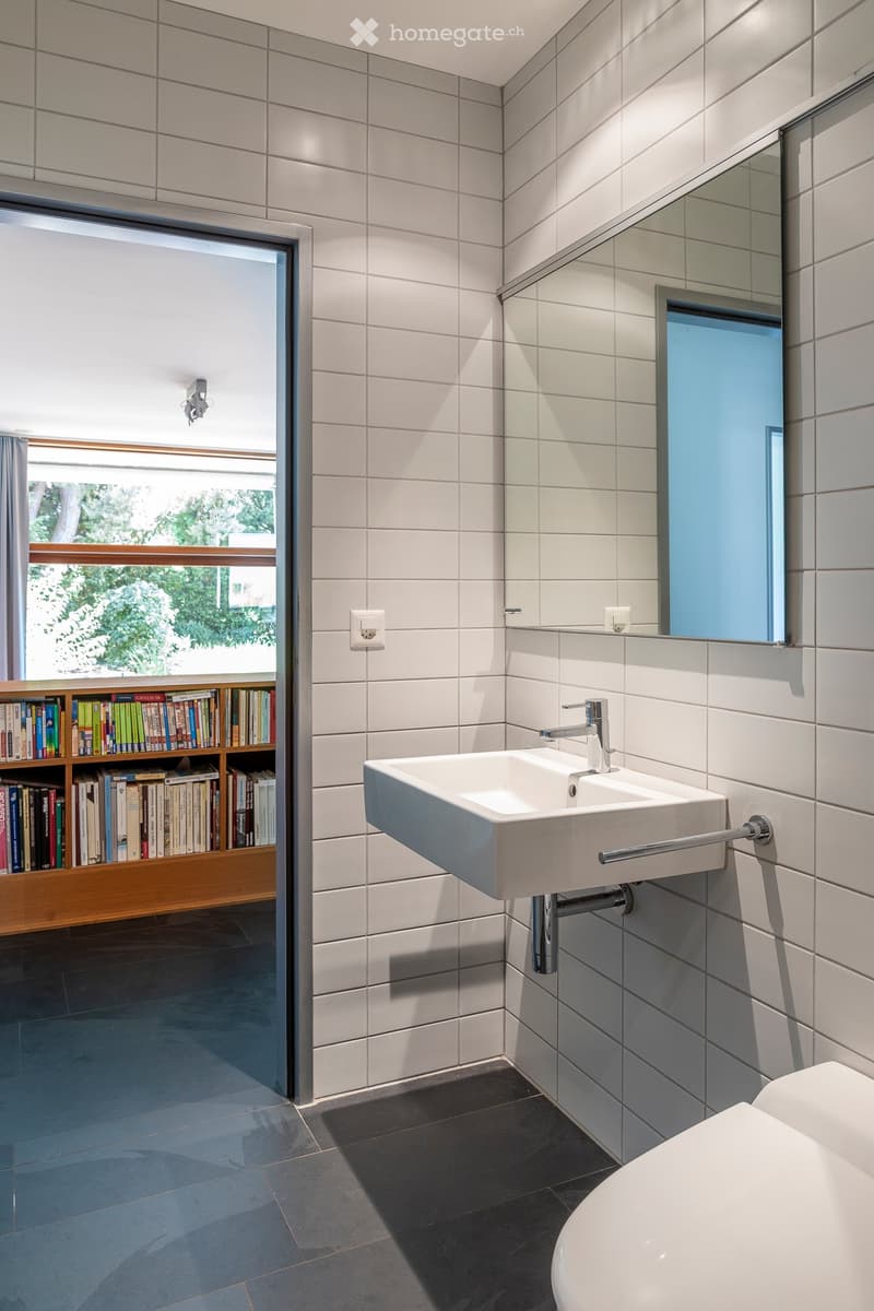 Modern bathroom with white tiles, a white sink with a chrome faucet, a toilet, a large mirror, and a bookshelf with books visible through an open doorway.