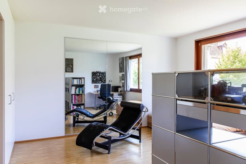Modern, minimalist room with light wooden flooring, large mirror, two black lounge chairs, a bookcase, a small table, and a cabinet with glass doors.