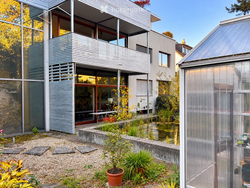 Modern house with a balcony, surrounded by a garden with a pond, potted plants, and a greenhouse.