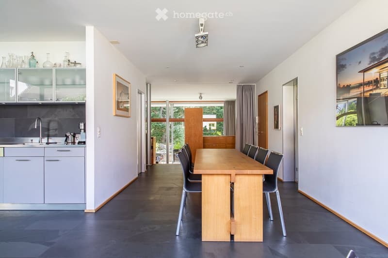 Modern kitchen and dining area with light grey cabinets, a wooden dining table, and six chairs. The kitchen features a sink, faucet, and coffee maker, with glassware displayed on top cabinets. Large windows provide natural light, and framed artwork adorns