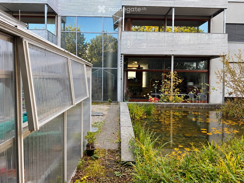 Modern house with a pond, greenery, and a greenhouse structure. The house features large glass windows and a sleek, contemporary design.
