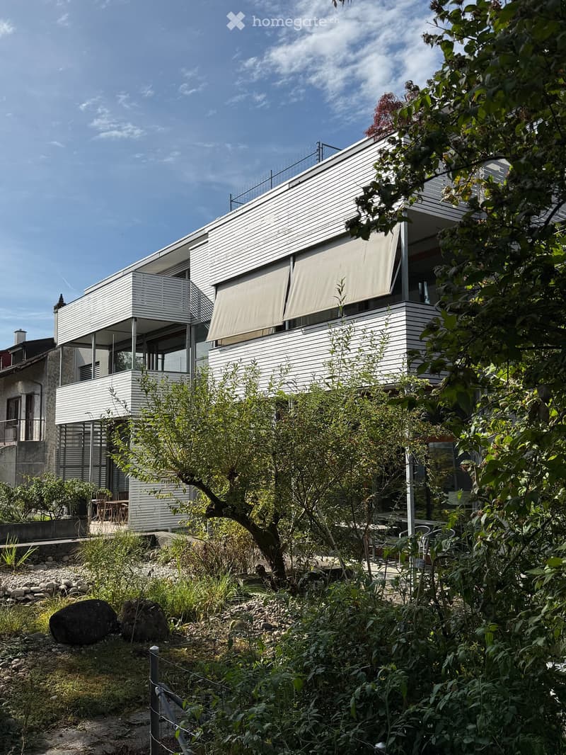 Modern two-story house with white siding, large windows, and a balcony. The surrounding area includes a garden with trees, rocks, and a small seating area.