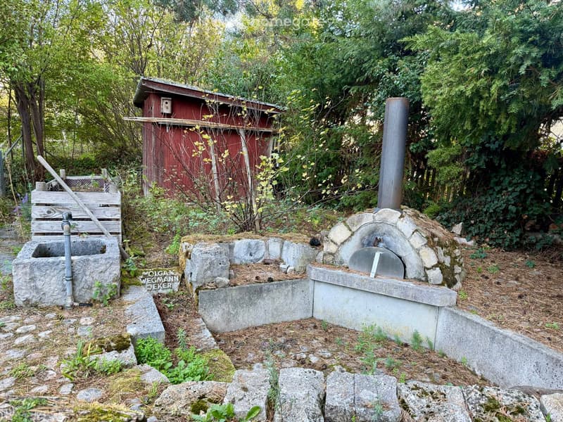Garden area with a stone well, wooden bench, red wooden shed, and stone oven with a chimney.