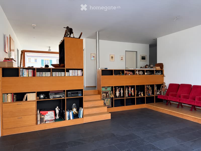 Modern living room with dark tiled floor, wooden bookshelf, red sofa, staircase leading to upper level, and various decorative items including sculptures and framed pictures.