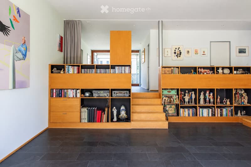 Modern living room with dark tiled floor, large wooden bookshelf filled with books, decorative items, and toys. A staircase leads upwards, with artwork and framed pictures on the walls.