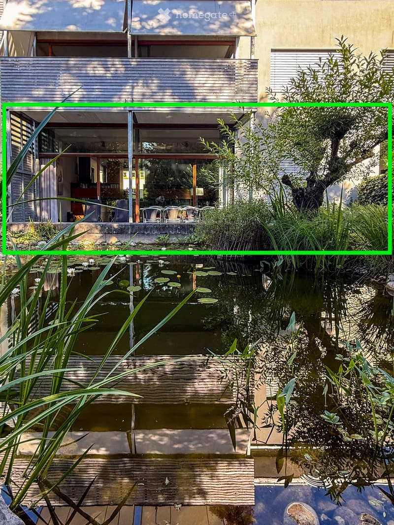 A modern outdoor area with a pond, lush greenery, and a covered seating area with chairs and a table.