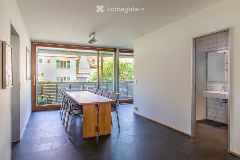 Modern dining area with a wooden dining table and chairs, a vase on the table, and a framed picture on the wall. The room has dark wooden flooring and white walls, with a doorway leading to a bathroom and a large window opening to a patio.