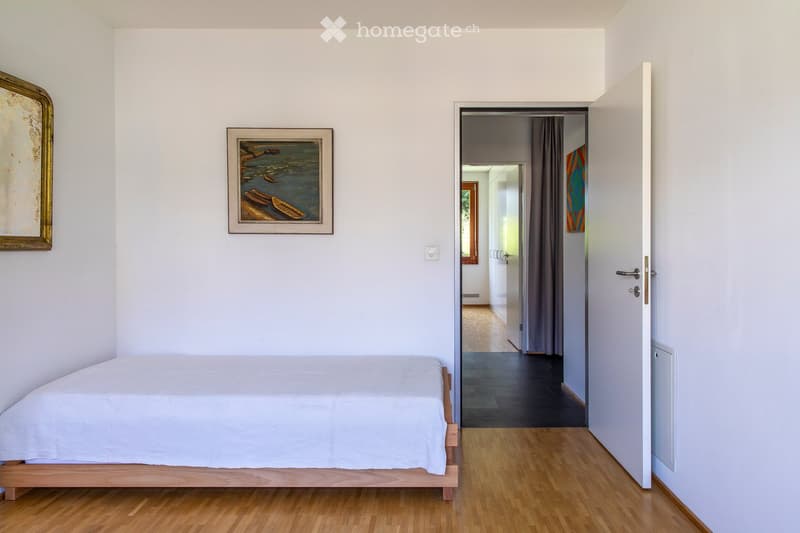 Minimalist bedroom with a single bed, light wooden flooring, and two framed pictures on the wall. An open doorway leads to a hallway with wooden flooring and a view of another room.