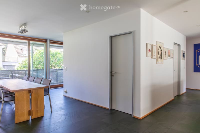 Modern dining area with a wooden table and chairs, large windows with a view of greenery, and artwork on the walls.