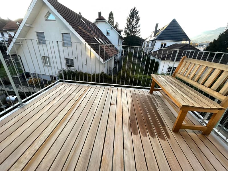 Wooden deck with a bench, metal railing, and view of neighboring houses and greenery.