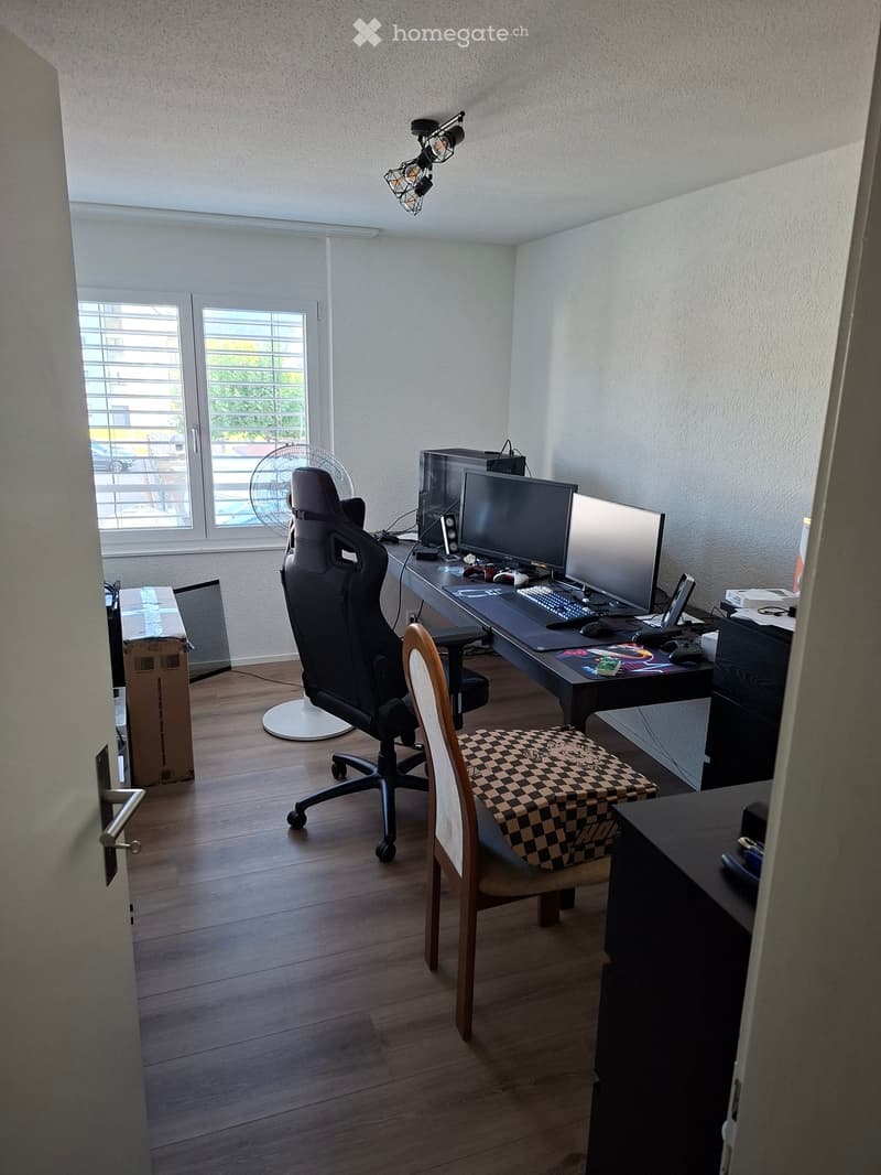 A well-lit room with wooden flooring, featuring a gaming chair, desk with dual monitors, keyboard, mouse, and a fan near the window. There is also a small table with a checkered cloth and a storage unit.
