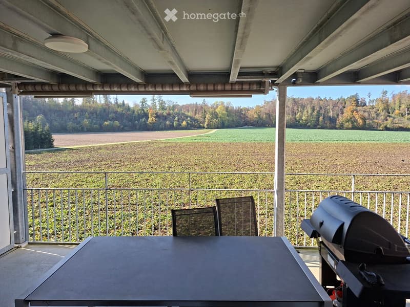 Balcony with a table, chairs, and a grill, overlooking a scenic countryside view.