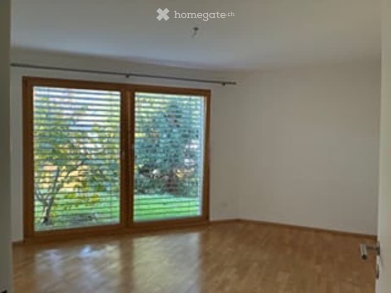 Empty room with wooden flooring, large sliding glass door leading to a green outdoor area, white walls, and minimal furnishings.