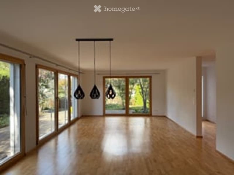 Bright, empty room with large windows and wooden flooring, featuring modern pendant lights and a view of greenery outside.