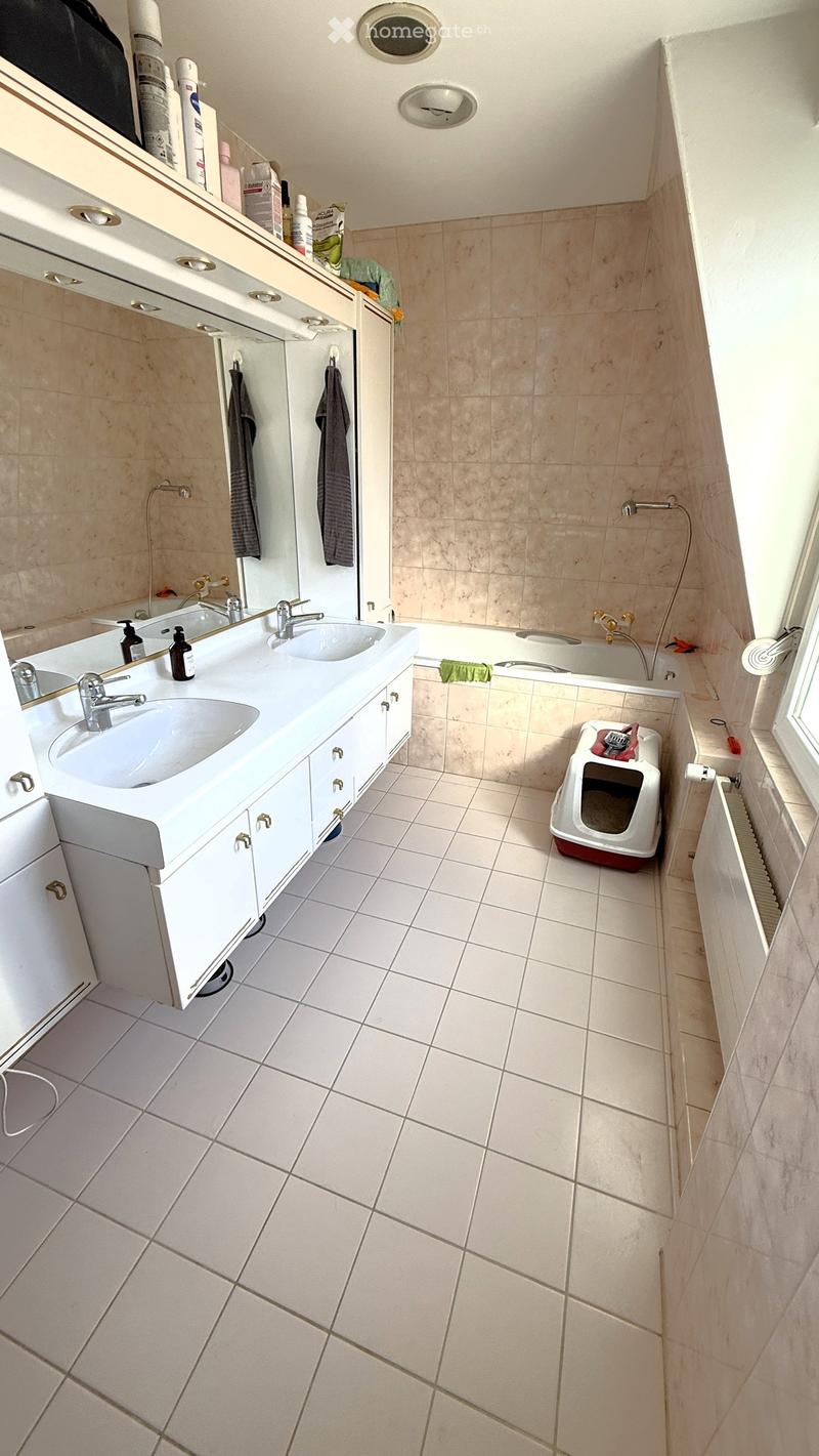 Bathroom with white tiled floor and walls, double sink vanity with mirrors, bathtub, and a cat litter box in the corner.