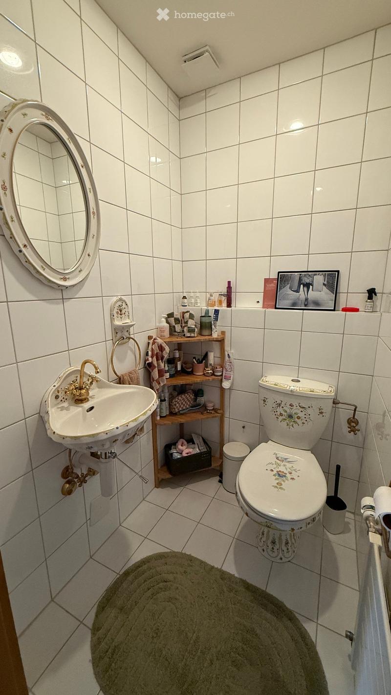 A small bathroom with white tiled walls and floor, featuring a sink with a gold faucet, a toilet with a floral design, a round mirror, a wooden shelf holding toiletries, a towel, a picture frame, and a green rug.