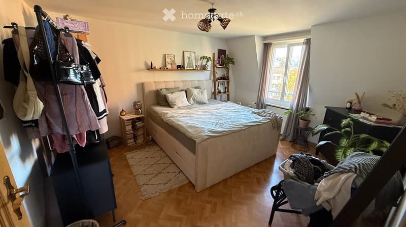 Cozy bedroom with a large bed, nightstand, chair, and various decorative items including plants and a shelf with books and pictures.