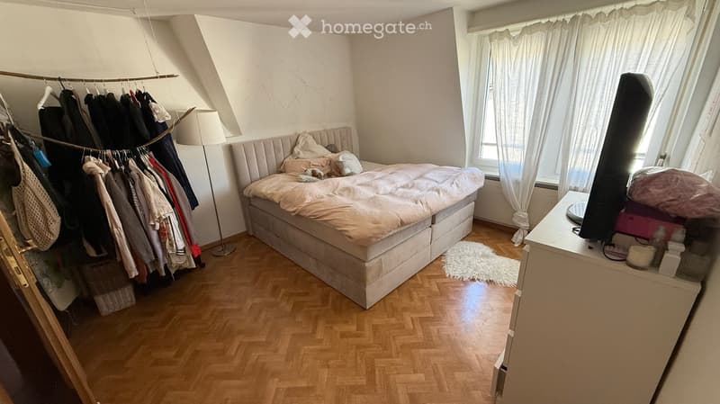 Cozy bedroom with a large bed, nightstand, lamp, clothing rack, and a mirror. The room features wooden flooring and large windows with white curtains.