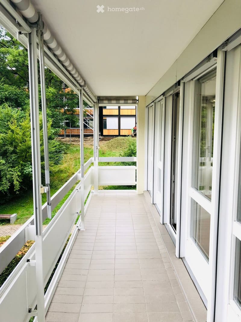 A modern, clean corridor with white tiled flooring, large windows on one side, and a white railing leading to a green outdoor area.