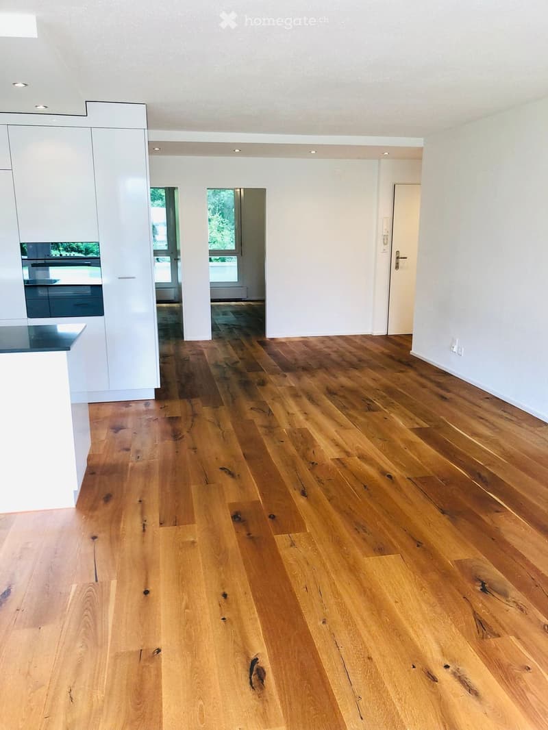 Empty room with light-colored wooden flooring, white walls, and recessed lighting. The room features a doorway and a glimpse of a kitchen area with white cabinets and a dark countertop.