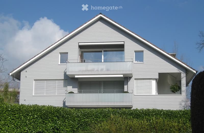 Modern two-story house with grey siding, large windows, and a balcony on the upper floor.