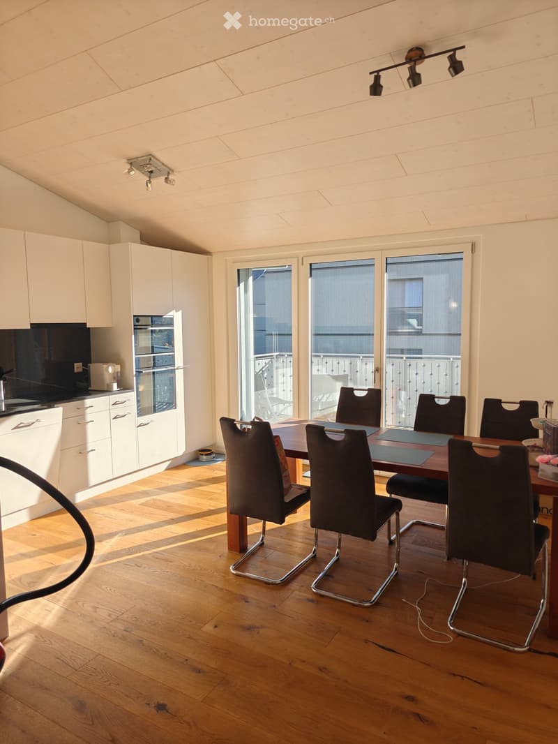 Bright, modern kitchen-dining area with wooden flooring, white cabinets, a wooden dining table with six black chairs, and large windows leading to a balcony.