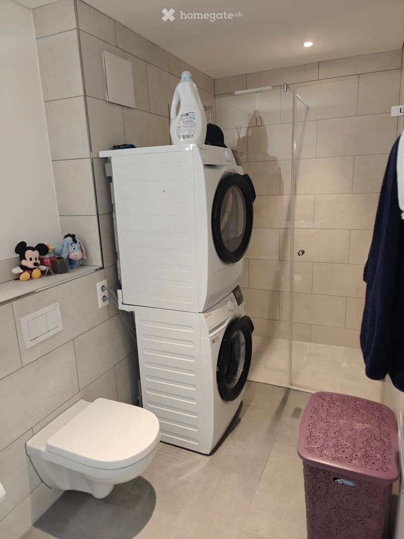 Modern bathroom with a toilet, stacked washing machine and dryer, laundry basket, and toiletries on the shelf.
