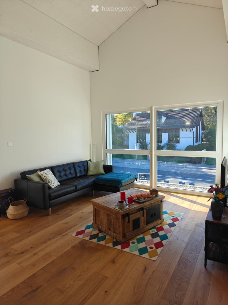 Modern living room with wooden flooring, large windows, a black leather sofa, a wooden coffee table with candles, a colorful rug, and a vase with flowers on a side table.