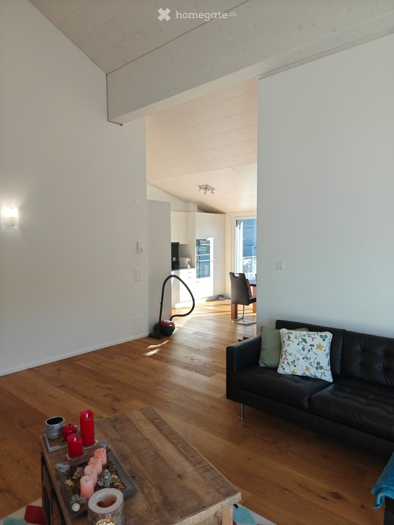Living room with wooden flooring, black leather sofa, coffee table with candles, and a chair in the background. The room has white walls and a slanted ceiling.