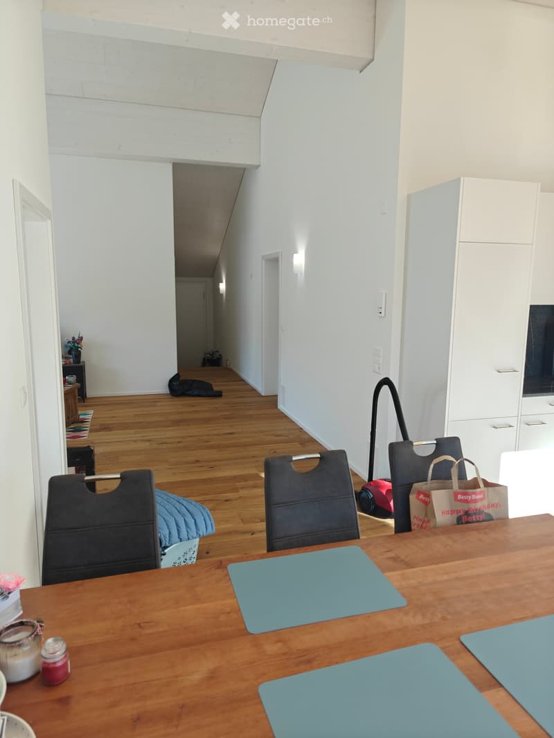 A modern, well-lit room with wooden flooring, a wooden dining table with chairs, a white cabinet, and a hallway leading to another room.