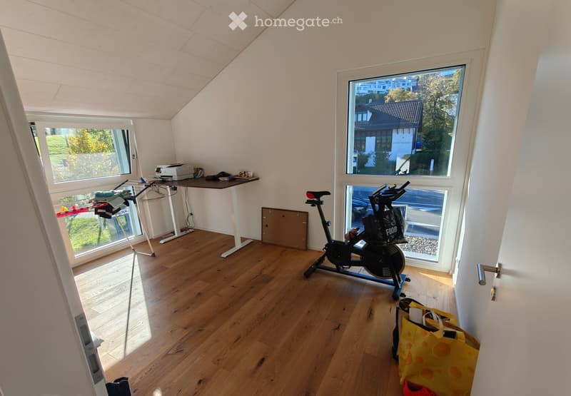 Bright room with wooden flooring, large windows, exercise bike, table with printer, and clothing rack.
