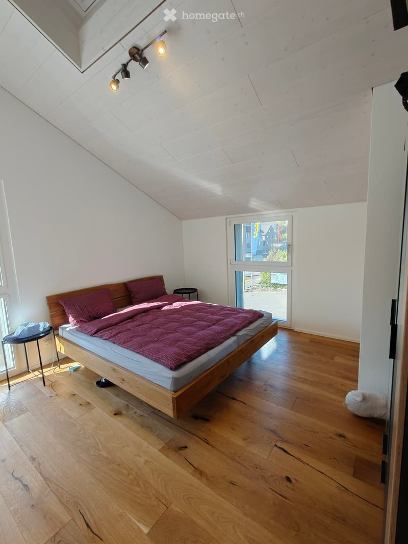 Bright bedroom with wooden floor, slanted ceiling, and large window offering a view. Features a wooden bed with red bedding, two small stools, and a stuffed toy on the floor.